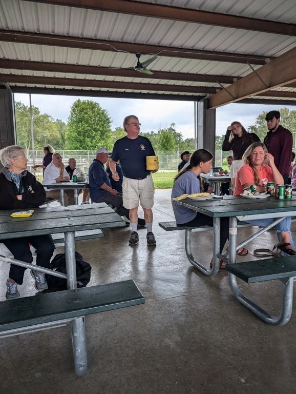 People seated at picnic tables in a covered pavilion, with one man standing in the center.