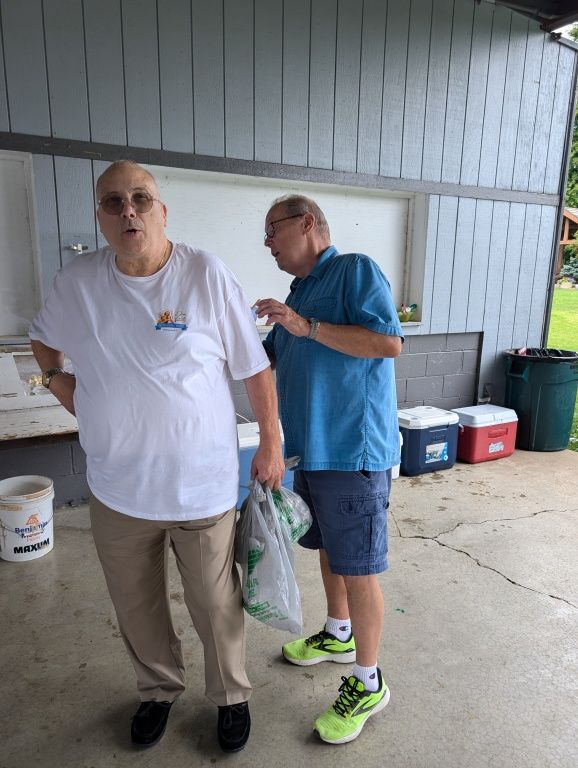 Two men standing under a covered outdoor area, one holding a bag and a drink, with coolers behind them.
