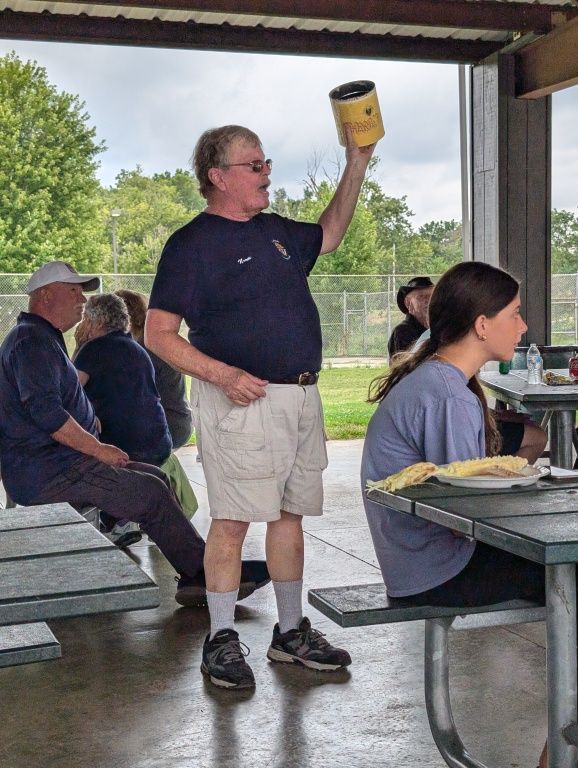 Man holding up a yellow container at a picnic shelter while others sit at tables nearby