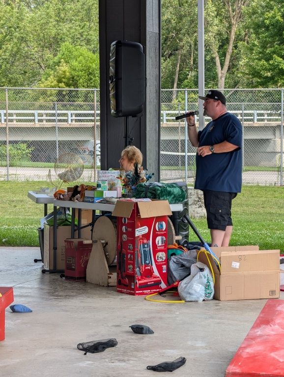 Person speaking beside a table of food and supplies under a pavilion, with a child seated nearby