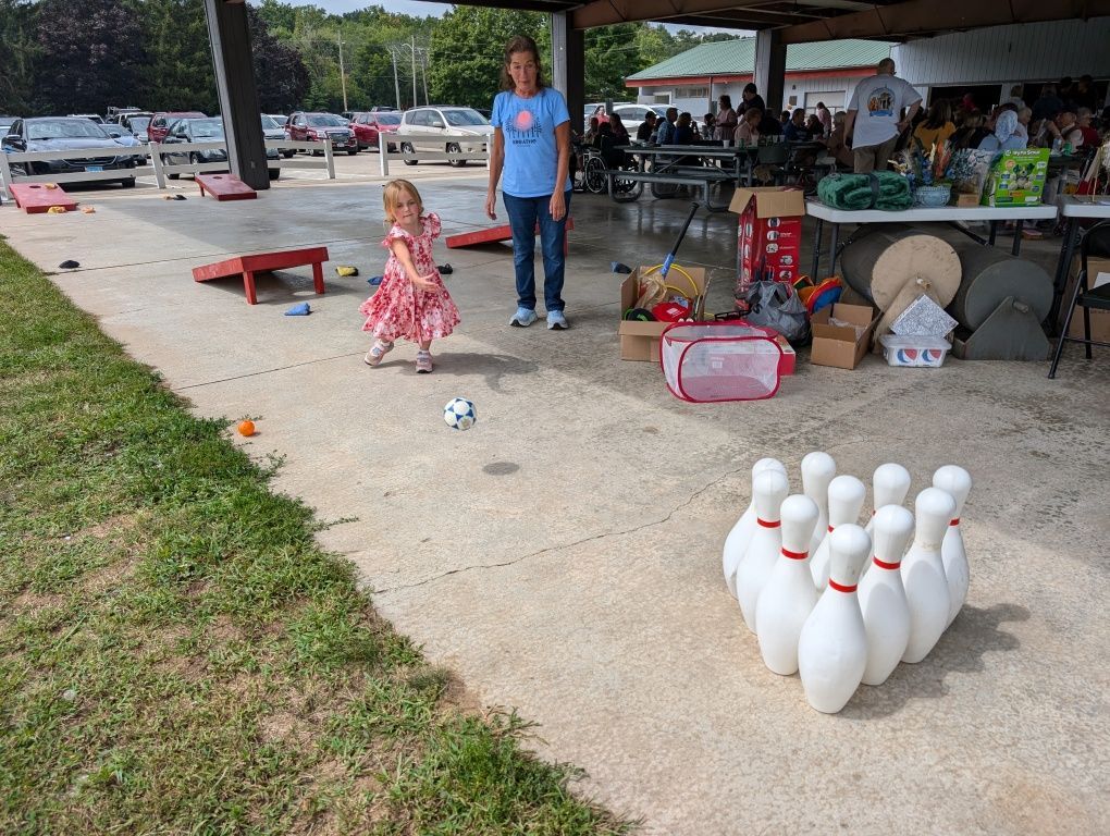 Child in pink dress near a bowling setup at a park pavilion, with pins and a ball on the concrete.
