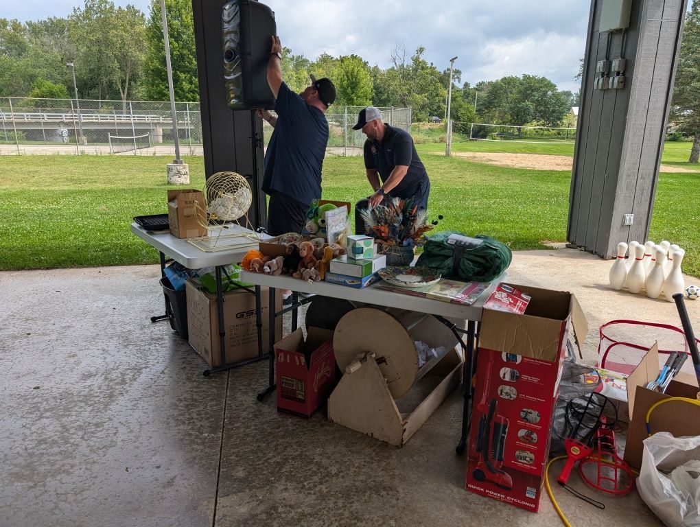 Two people at a picnic table under a pavilion, setting up items beside a grassy park field.