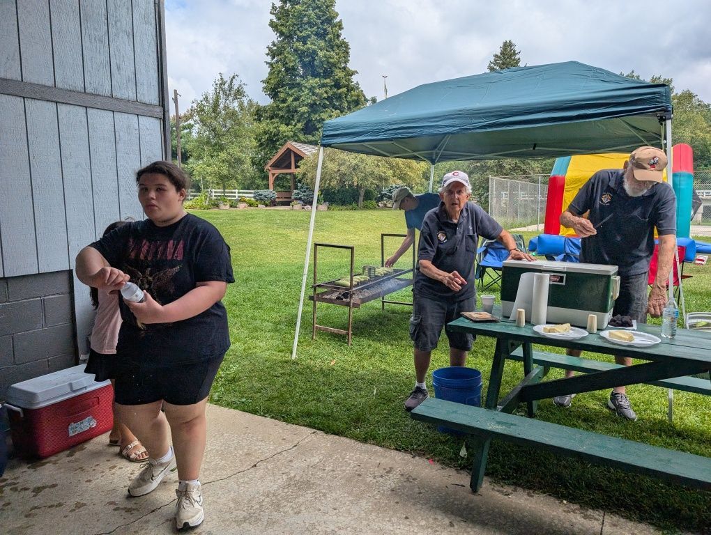 People under a green canopy at a backyard picnic, with folding tables, cooler, and yard games nearby