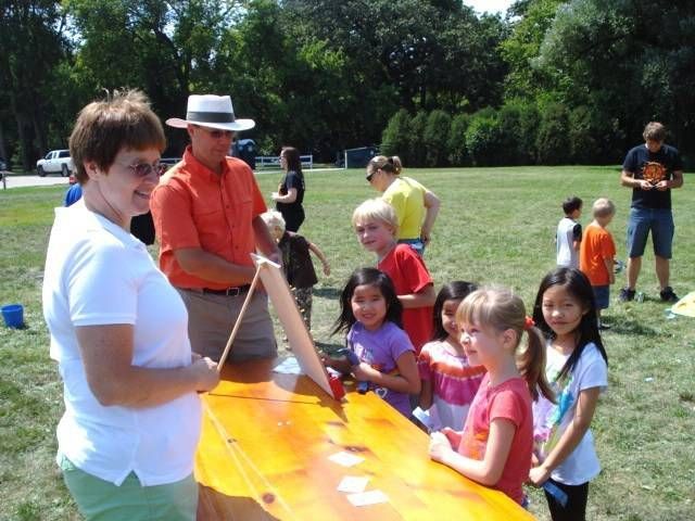 Adults and children hold a bright orange kite in a sunny park.
