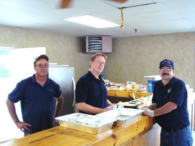 Three people stand behind a food table in a room with trays and boxes of prepared food.
