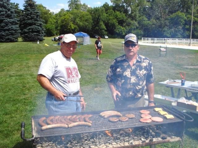 Two men grilling sausages and burgers at a smoky outdoor picnic