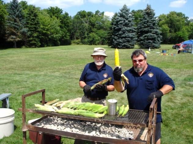 Two people grilling food outdoors at a park picnic table under sunny skies