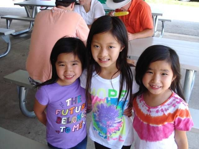 Three smiling children standing together at a picnic table outdoors