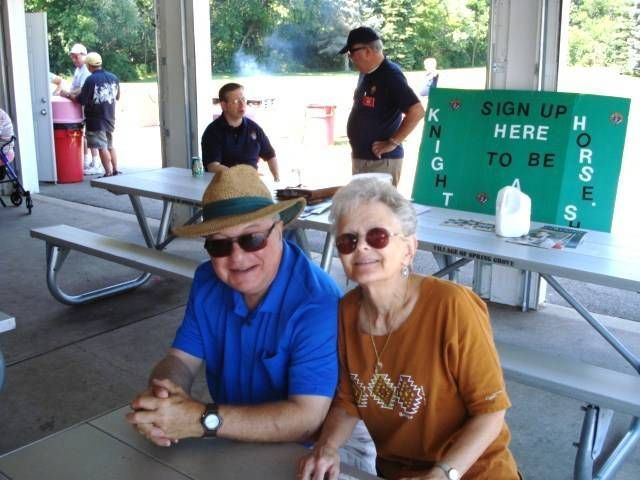 Two people seated at a picnic table in a park pavilion, with a sign reading “Sign up here to hose” nearby.