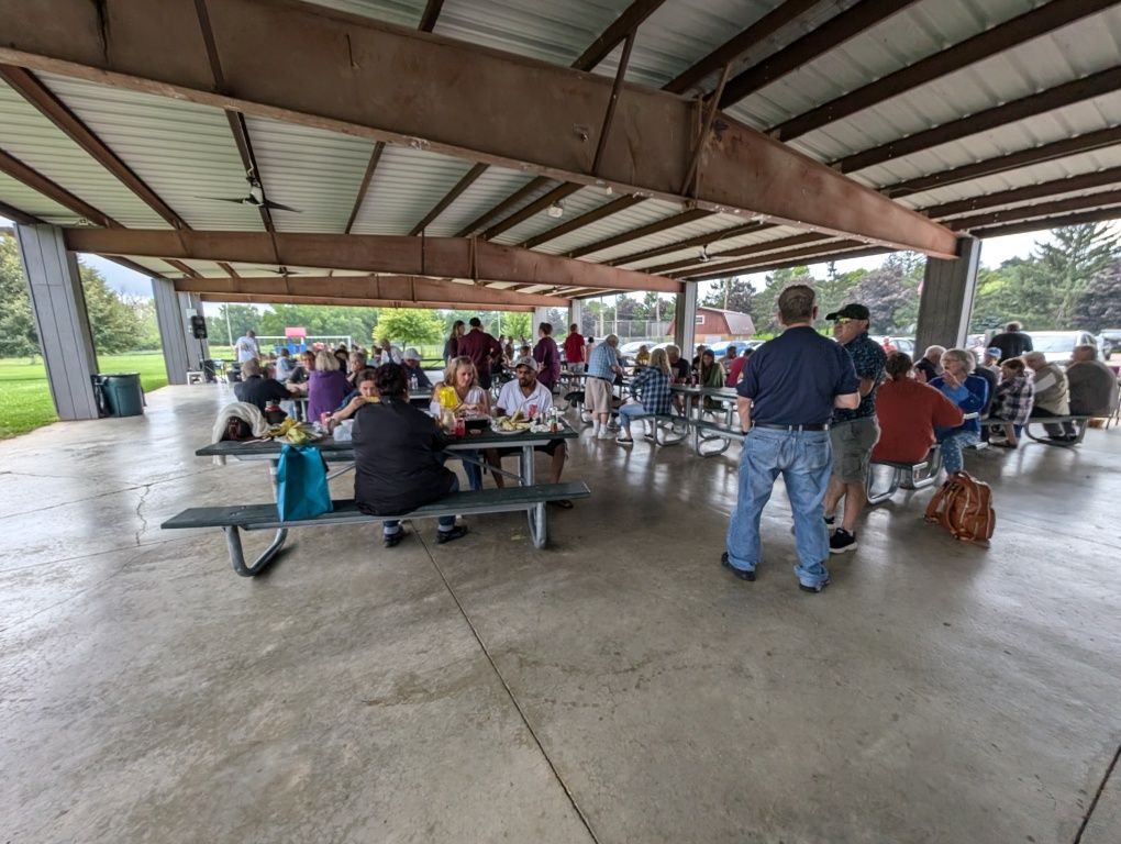 People gathered under a pavilion at picnic tables in a park, with dogs nearby on a wet concrete floor.