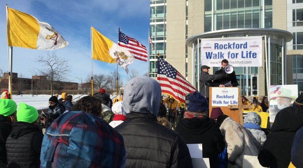 Crowd holding signs and flags at a rally outside a building, with a “Rockford Walk for Life” banner.