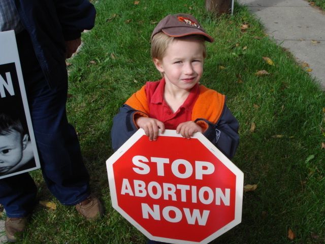 Child holding red STOP ABORTION NOW sign outdoors on grass