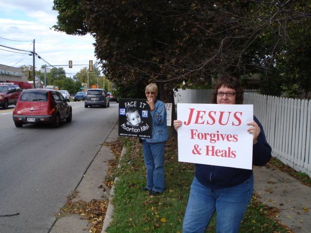 Two protesters on a roadside holding signs: “JESUS Forgives & Heals” and “HE IS REAL.”