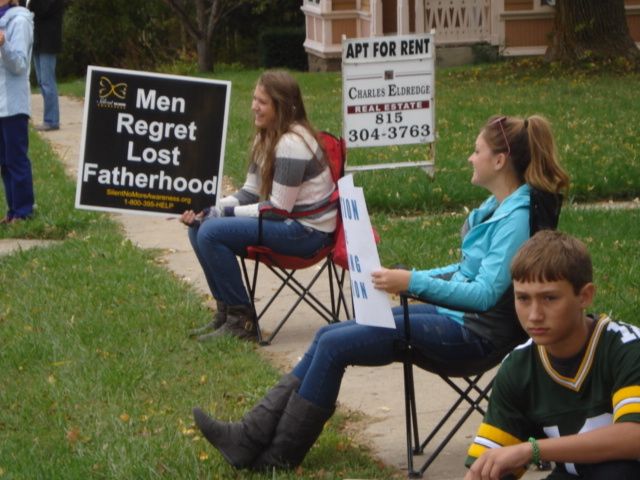 People sitting on folding chairs hold protest signs on a grassy lawn, including “Men Regret Lost Fatherhood.”