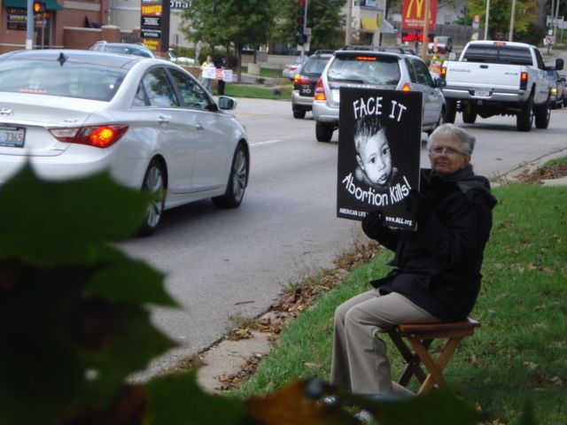 Person seated on the curb holds a protest sign beside a busy street with passing cars.