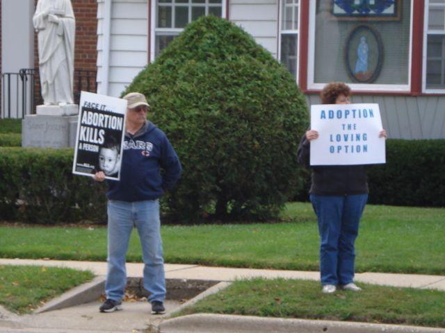 Two protesters stand outside a building holding anti-abortion signs.