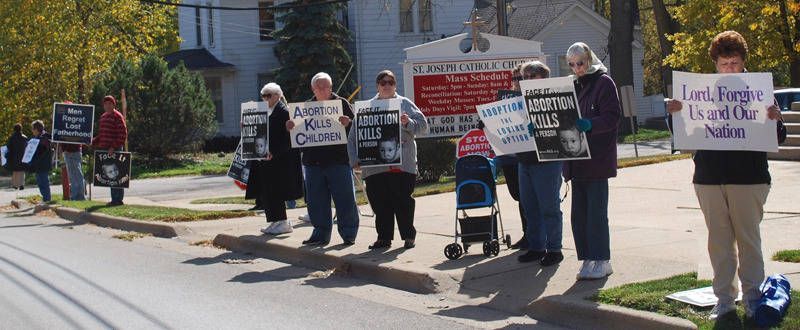 People holding protest signs line a sidewalk in a residential neighborhood.