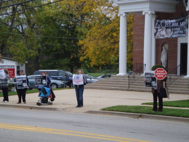Protesters holding signs outside a brick building with columns, one seated in a wheelchair.