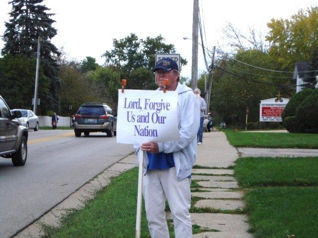 Man holding a sign on a sidewalk near a road that reads “Lord Forgive Us and Our Nation”