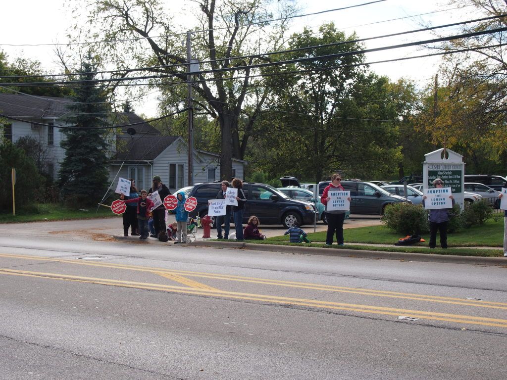 People holding signs and gathering along a roadside near parked cars and a residential building
