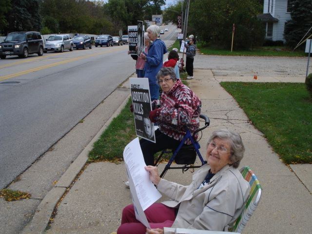 People seated in folding chairs along a sidewalk near a street, with signs and a grassy lawn nearby.