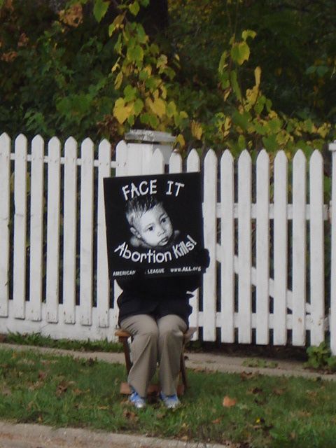Person in front of a white picket fence holding a black sign that says “Face It Abortion Kills.”