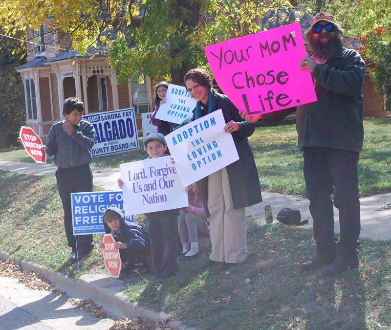 Protesters holding signs on a sidewalk, including “Your Mom Chose Life” and “Voted for Republican, Free the People”