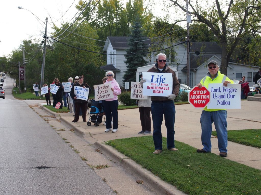 People holding protest signs along a suburban roadside, including one reading “STOP the killing NOW.”