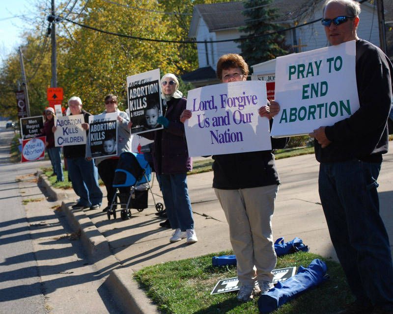 Pro-life protestors holding signs on a sunny sidewalk, including “PRAY TO END ABORTION.”