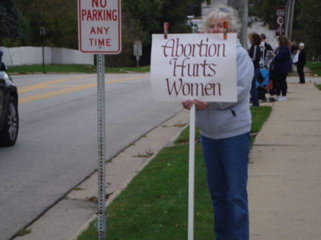Protester on sidewalk holding sign reading “Abortion Hurts Women” near a no-parking sign.