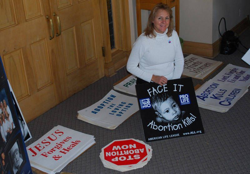 Woman kneels beside protest signs on carpet, including “Face It” and “Abortion” messages.