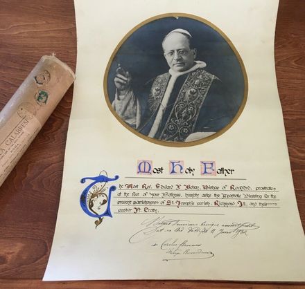 Formal certificate with a portrait seal and ornate calligraphy on cream paper, beside a rolled document on wood.