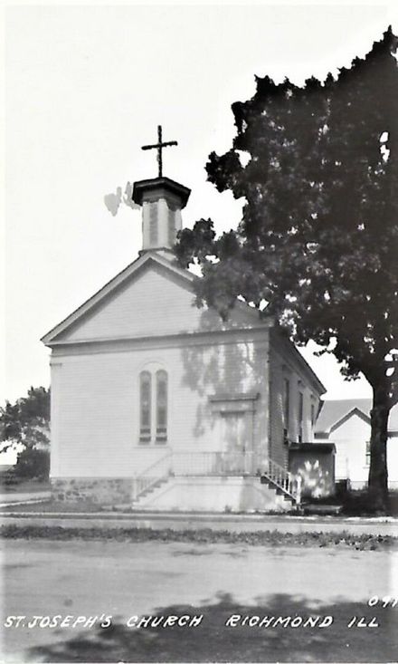 Small white church with steeple and cross beside a large tree, in a quiet rural setting.