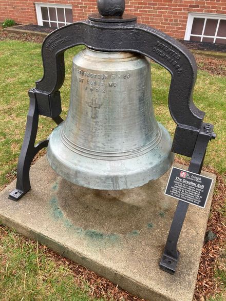 Large historic metal bell mounted on a concrete base beside a brick building.