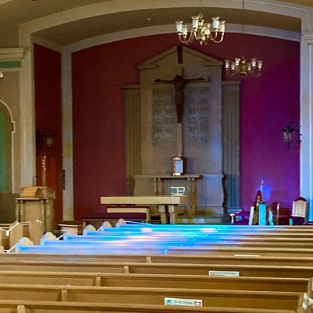 Church sanctuary with wooden pews, altar, and large cross on the front wall, lit by chandeliers.