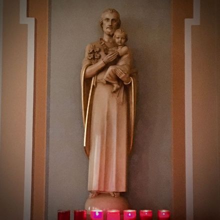 Statue of a saint holding a child, with lit candles below, in a church alcove.