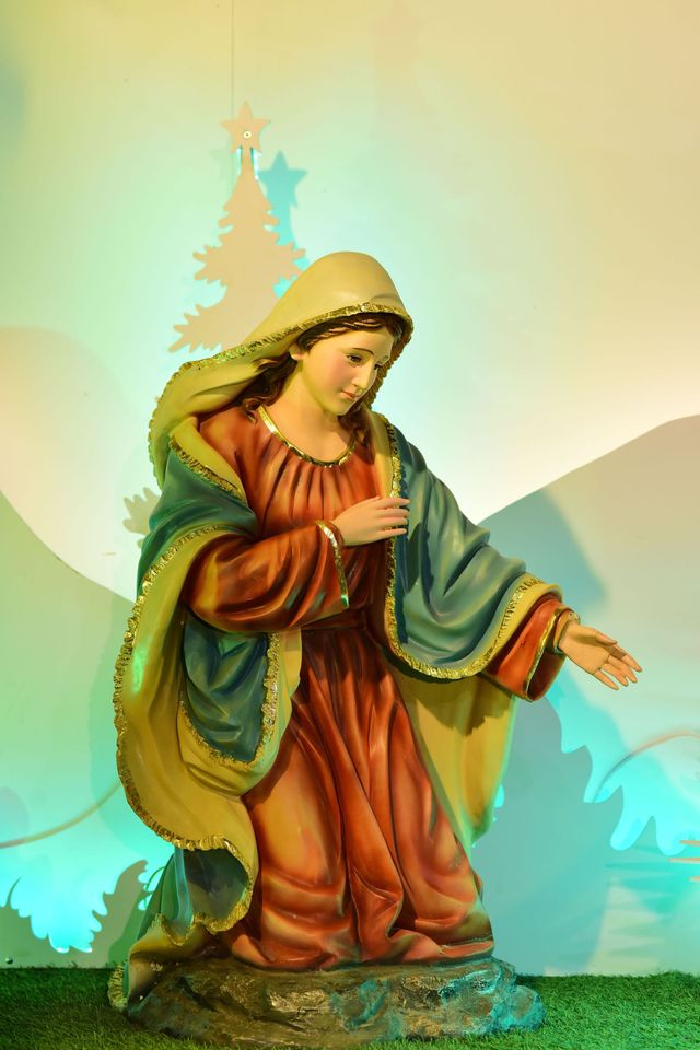 Nativity figurine of a robed woman reaching out, with a pastel sky and silhouetted trees behind her