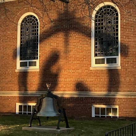 Large bell statue on a lawn in front of a red brick building with arched windows and shadows