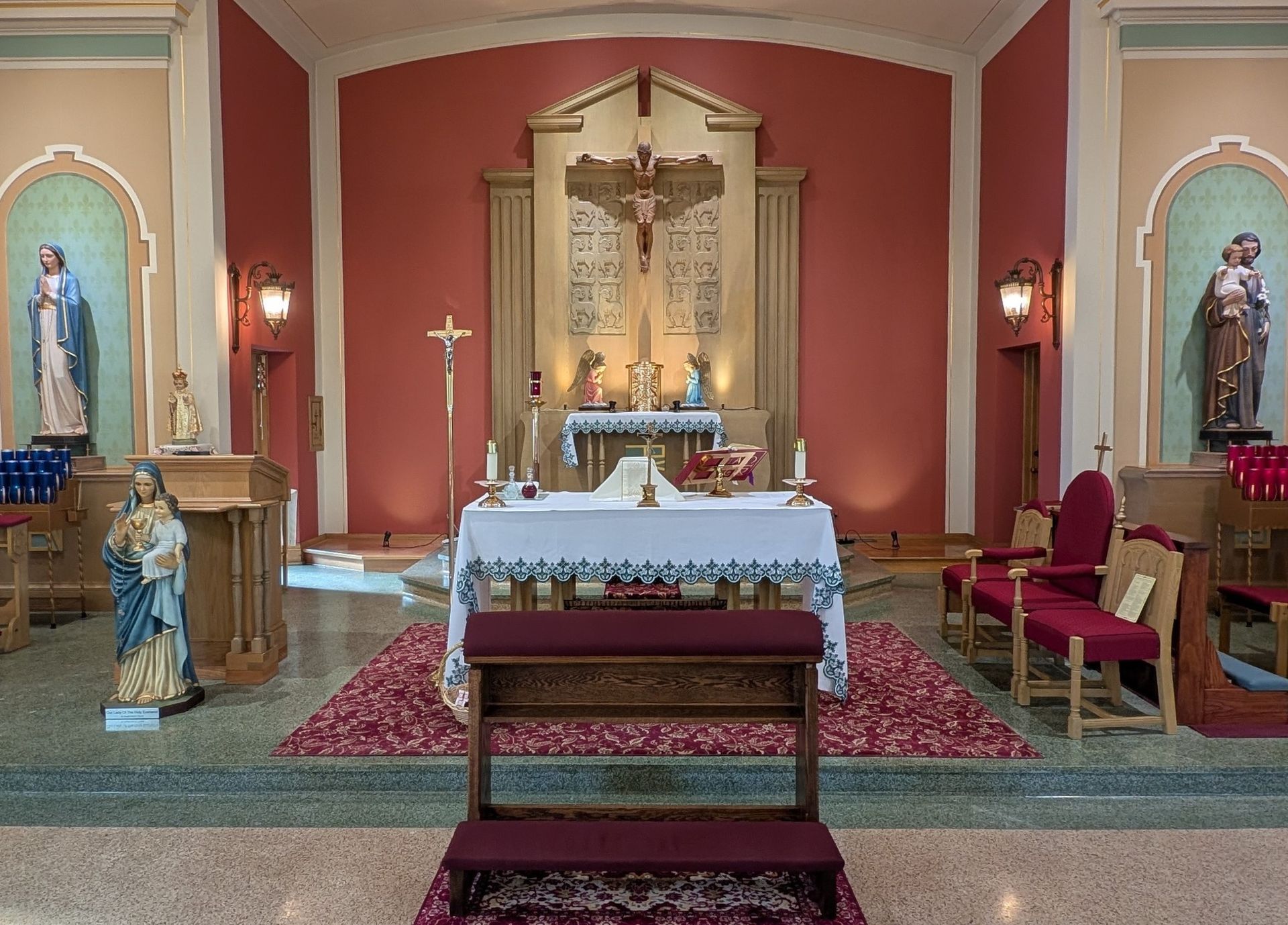 Church altar with crucifix, white cloth, candles, statues, and red walls in a chapel sanctuary