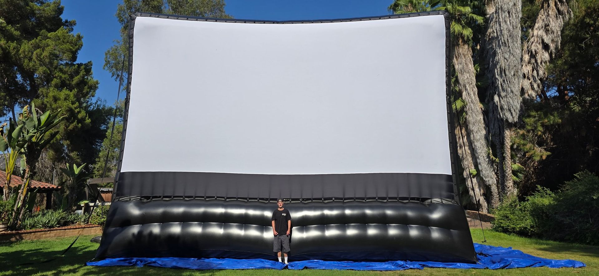An inflatable outdoor movie screen set up in a grassy field; a person stands next to it. A treeline is in the background.