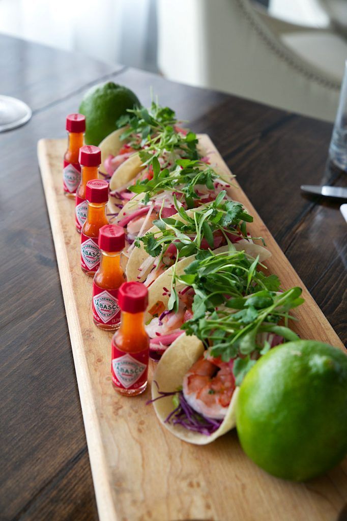 A long wooden cutting board topped with tacos and bottles of tabasco sauce.