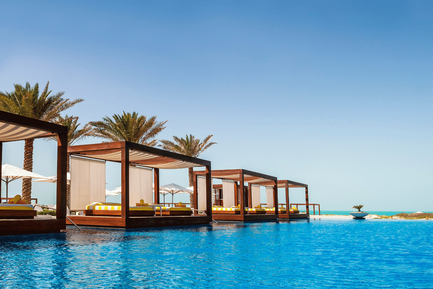 Luxury poolside cabanas beside a bright blue infinity pool under palm trees and clear sky