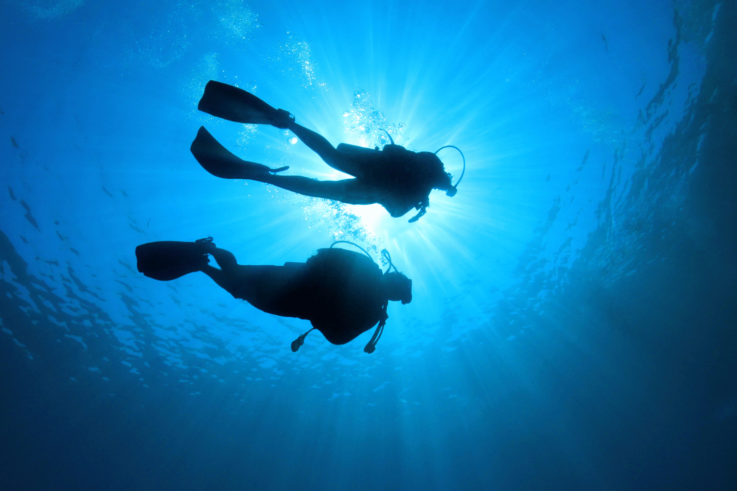 Two scuba divers silhouetted in blue underwater light, swimming beneath the surface