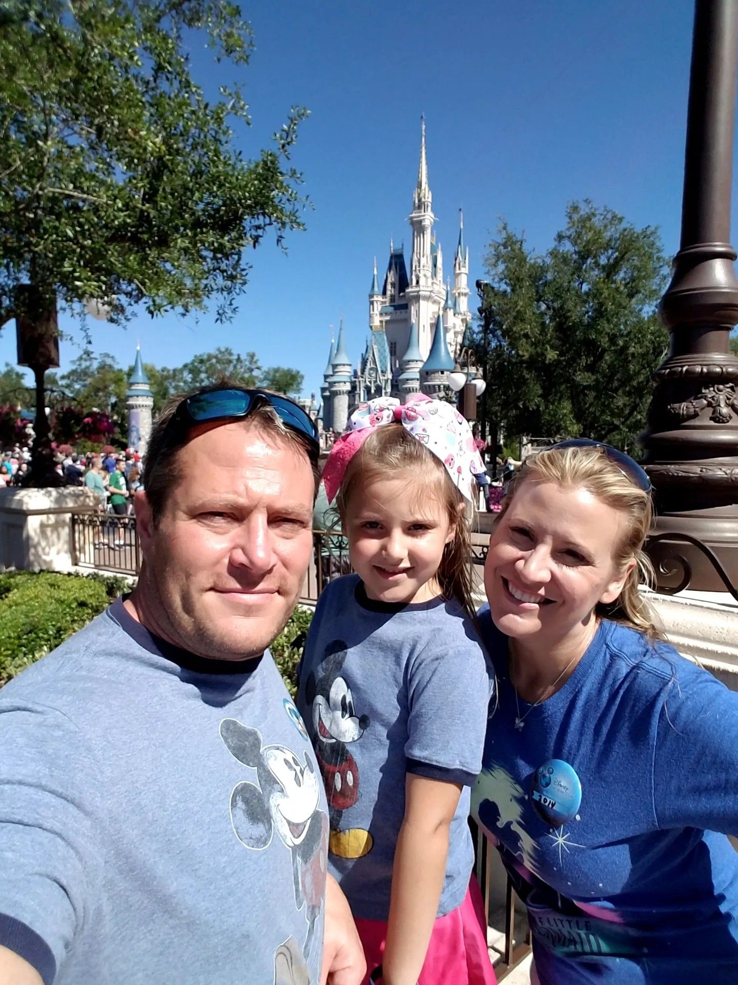 Three people smiling in front of a castle at a theme park on a sunny day.