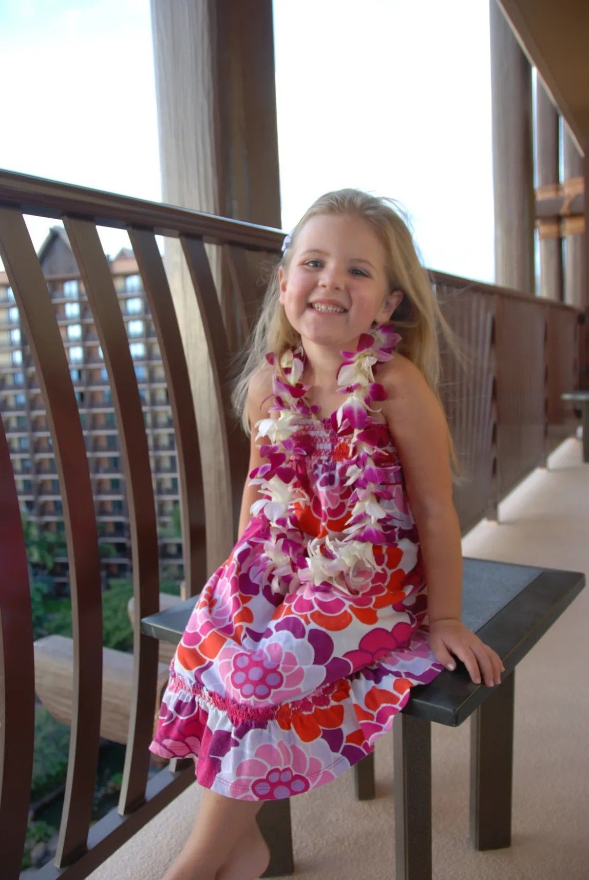 Smiling child in a colorful dress sitting on a balcony chair with city buildings in the background
