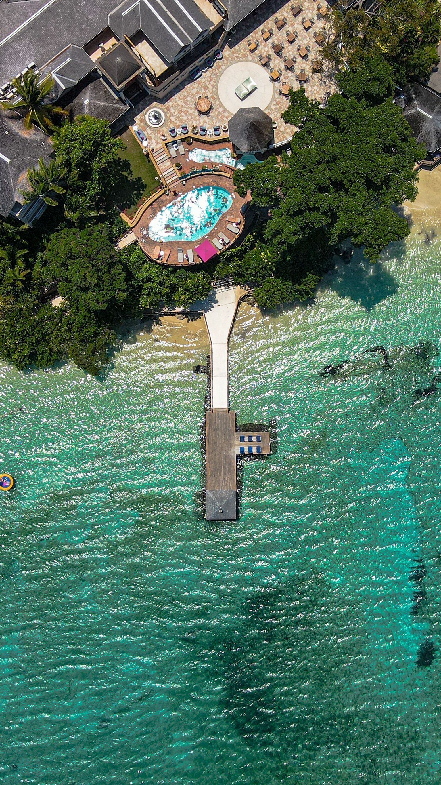 Aerial view of a turquoise beach cove with a small dock, pool, and palm trees beside rocky shore.