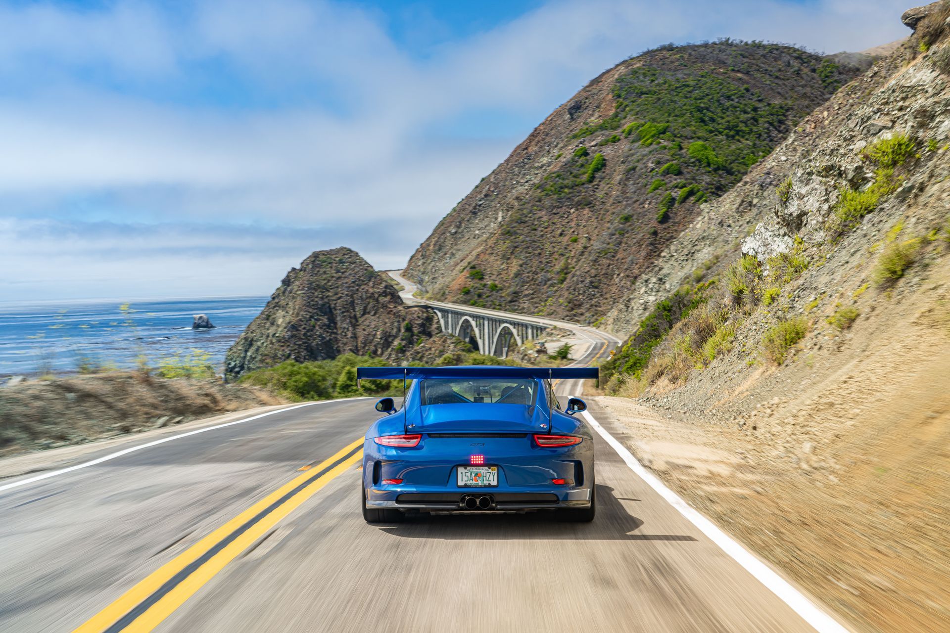 Blue sports car driving on a coastal highway beside cliffs and the ocean
