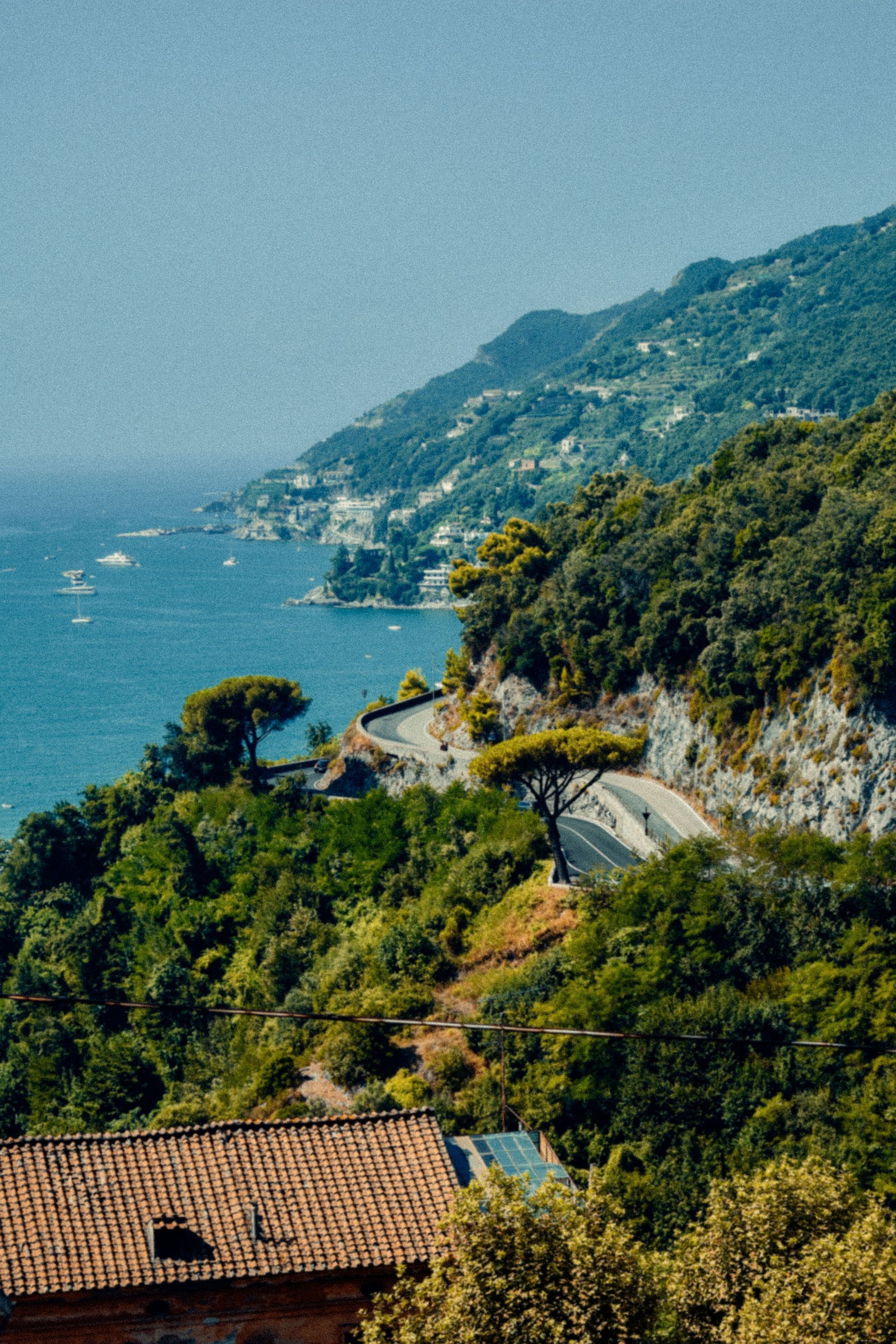 Coastal hillside road above blue sea, with terracotta roofs and green cliffs in the foreground