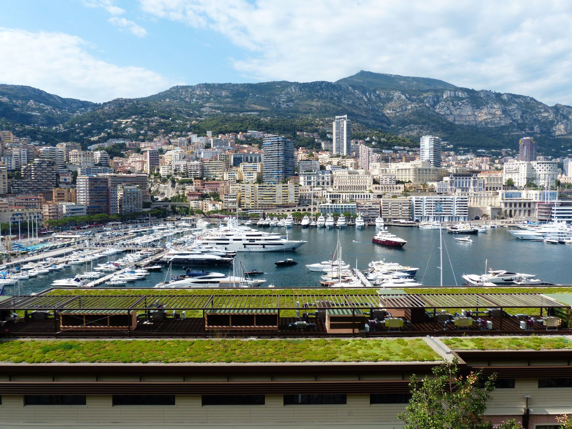 Marina with yachts and sailboats in a bay, backed by a dense coastal city and mountains under a clear sky