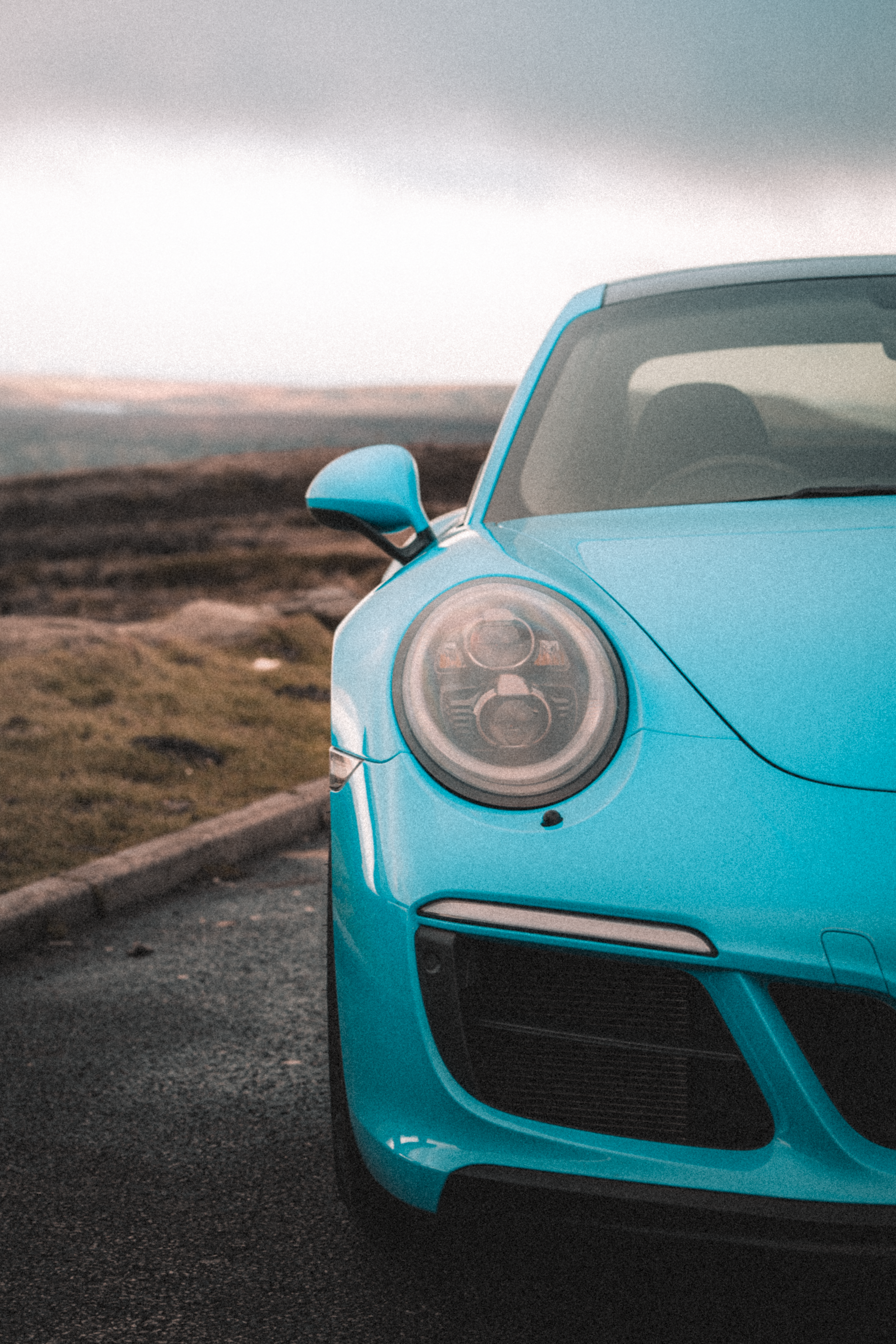 Turquoise sports car parked on a road, close-up of the front left headlight at dusk.